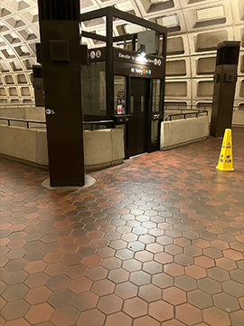 A picture of an elevator. To the left and right in the background of the image there are two wayfinding structures. To the left of the image there is a yellow wet floor cone. In the center of the background is the elevator, with a light shining down to illuminate the typeface on its surface.